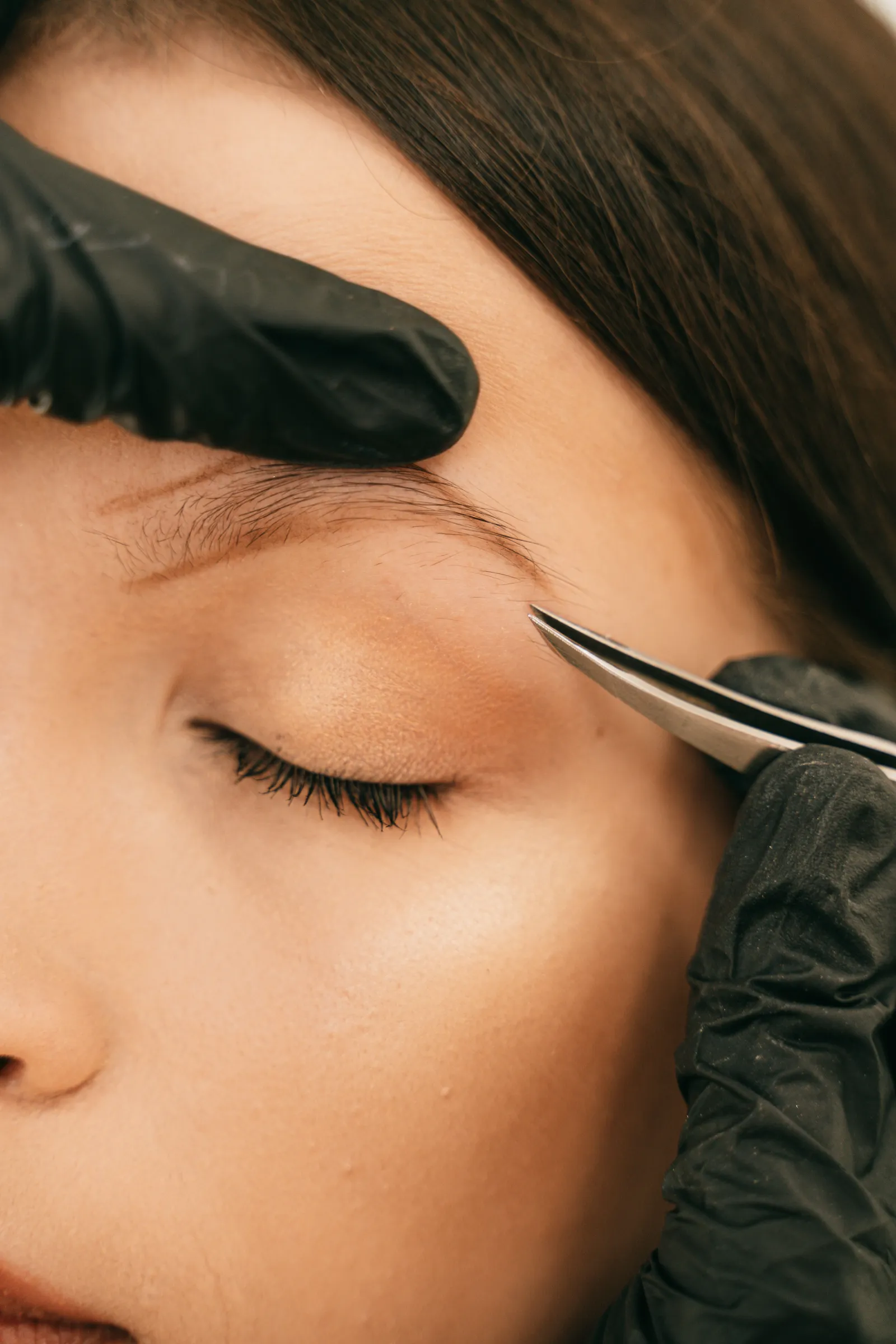 Primer plano del ojo cerrado de una clienta mientras la esteticista, con guantes negros, tensa la piel de la frente con un dedo y extrae un pelo del contorno de la ceja con unas pinzas de punta fina