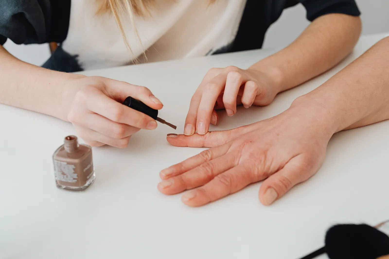 Manos de una esteticista aplicando esmalte color nude en la uña del dedo corazón de una clienta; las dos manos se apoyan en una mesa blanca, con el frasco de esmalte al lado