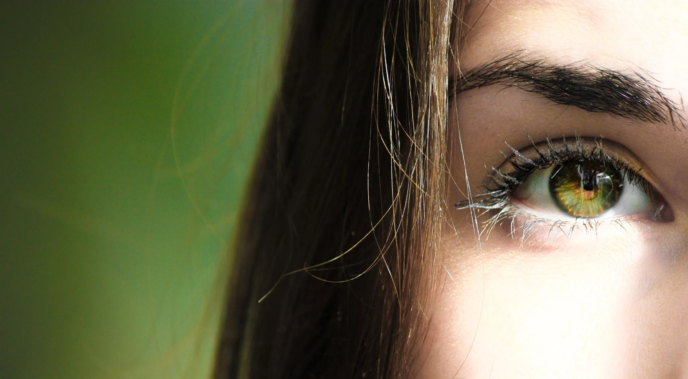 Primer plano del ojo de una mujer joven mirando hacia arriba, con la ceja depilada y definida y el iris verde iluminado por la luz del sol; el pelo castaño le cae sobre la sien y el fondo queda desenfocado en verde